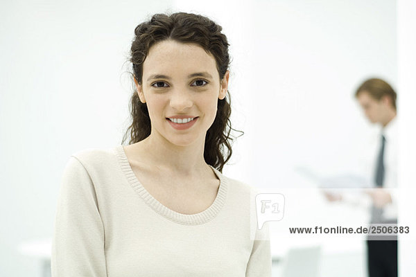 Junge Berufsfrau lächelt vor der Kamera  Kollegin im Hintergrund