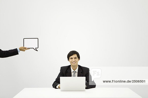 Young businessman sitting with laptop  smiling at camera  colleague holding up blank word bubble