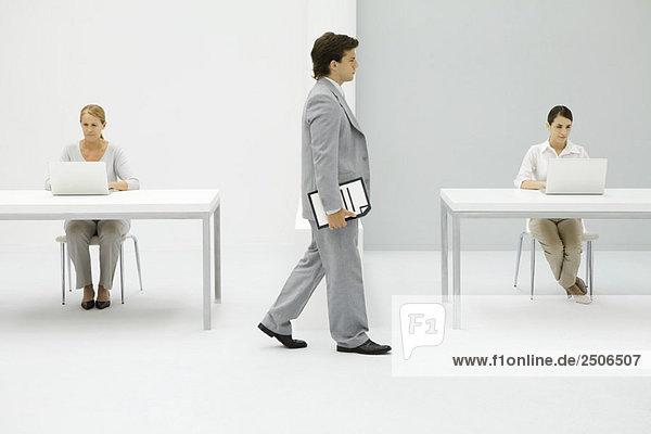 Businessman walking in office  carrying document under arm  female colleagues working in background