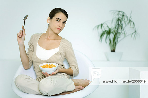 Young woman sitting in chair  holding cereal bowl and spoon  smiling at camera