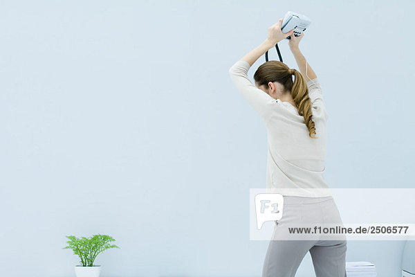 Junge Frau hält Festnetztelefon über dem Kopf bereit  um es am Boden zu zertrümmern  Rückansicht