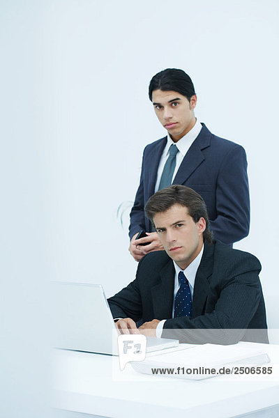 Two serious businessmen looking at camera  one sitting at desk  one standing behind him