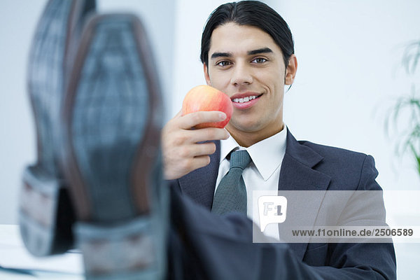 Young professional man sitting with feet up  holding apple and smiling at camera