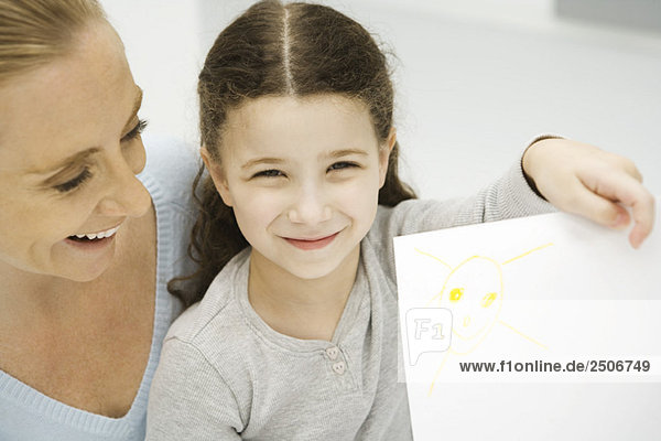 Girl holding up and showing drawing of the sun  mother smiling behind her daughter