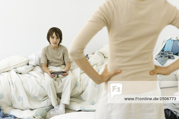 Junge sitzt im unordentlichen Schlafzimmer  hält Handheld-Videospiel  Mutter im Vordergrund mit Händen auf den Hüften  abgeschnitten