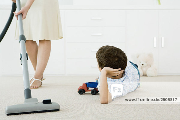 Junge  der mit Spielzeug auf dem Boden liegt und zu seiner Mutter aufblickt  die um ihn herum Staub saugt.