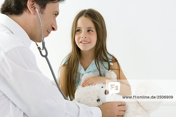 Kinderarzt  der vorgibt  den Teddybär eines Mädchens mit Stethoskop zu untersuchen.