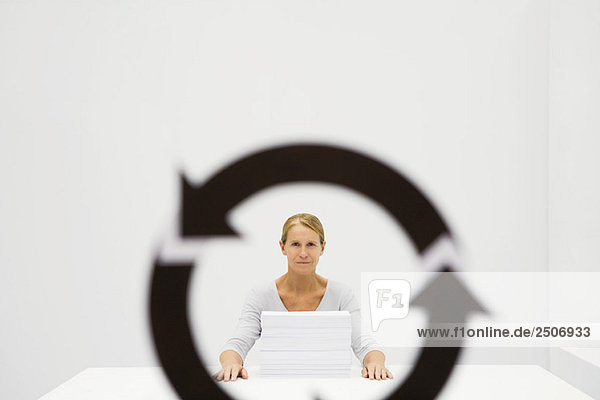 Professionelle Frau  die hinter einem Papierstapel sitzt und die Kamera durch das Recycling-Symbol betrachtet.