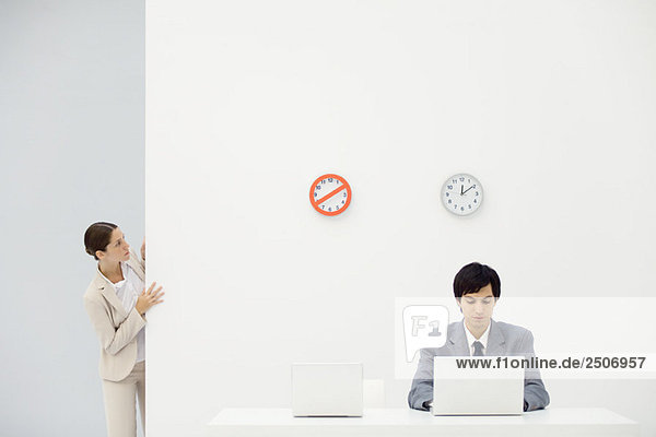 Büroangestellte schaut um die Ecke auf Uhren an der Wand  Kollegin blickt auf Laptop