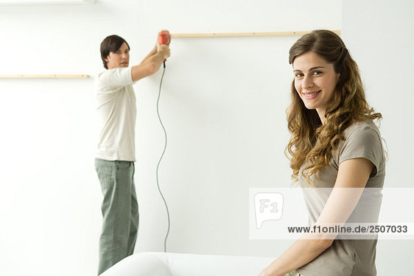 Man attaching thin piece of wood to wall  woman in foreground smiling at camera