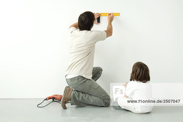 Mann  der die Wand mit einem Lineal misst  während sein Sohn zusieht  bohrt auf dem Boden in der Nähe.