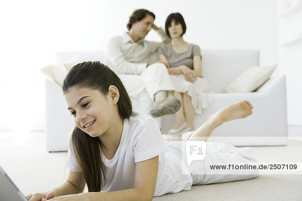Girl lying on stomach on the floor  parents sitting on couch in background