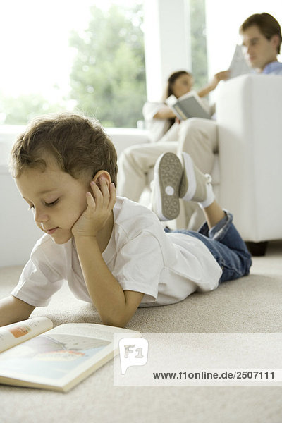 Kleiner Junge auf dem Boden liegend und Lesebuch  Eltern lesen auf Sofa im Hintergrund