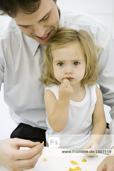 Young girl eating a piece of cereal  sitting on her father's lap  man helping her