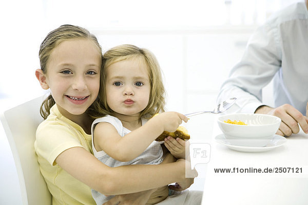 Junges Mädchen sitzt auf dem Schoß ihrer älteren Schwester  hält Löffel und Croissant und lächelt in die Kamera.