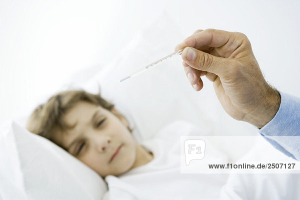 Kleiner Junge im Bett liegend  Männerhand mit Thermometer im Vordergrund