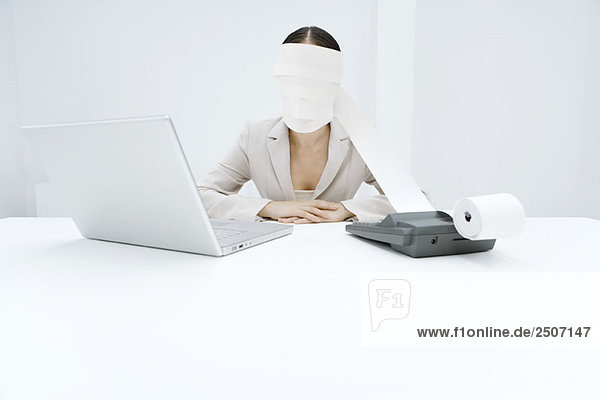 Frau am Schreibtisch sitzend mit Addiermaschine und Laptop  Ausdruckband um den Kopf gewickelt