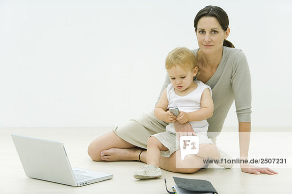 Professional woman sitting on the ground with laptop  holding toddler on lap  toddler looking at cell phone