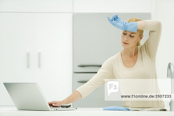 Frau steht am Küchenspülbecken  greift nach dem Laptop  trägt Gummihandschuhe