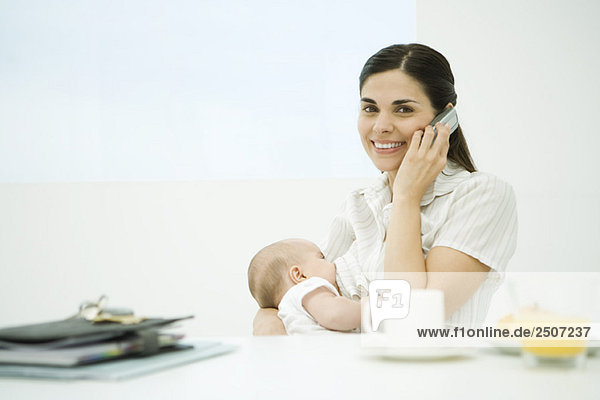 Frau am Frühstückstisch sitzend  Baby stillen  Handy benutzen  Kamera lächelnd
