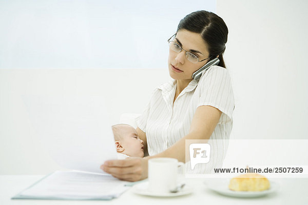 Professional woman sitting at breakfast table  holding baby  using cell phone and studying document