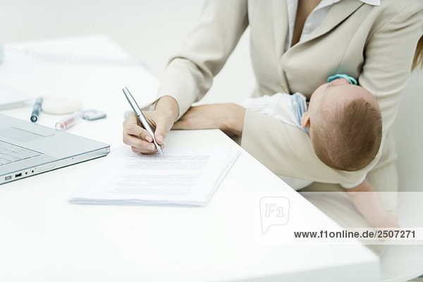 Professionelle Frau signiert Dokument am Schreibtisch  hält schlafendes Kind in der Hand  Ausschnittansicht