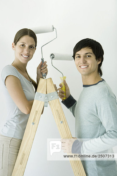 Couple standing on ladder  both holding up paint rollers and smiling at camera