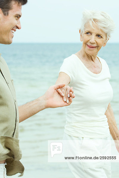 Senior woman walking hand in hand with adult son by water  smiling at each other