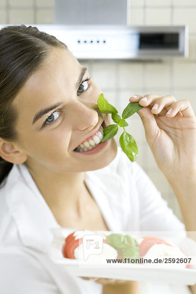 Woman biting into sprig of basil  holding plate of Caprese salad