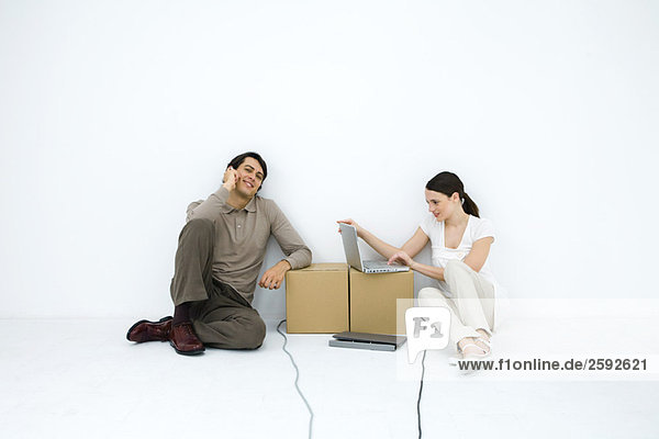 Man talking on phone  sitting on floor next to woman using laptop computer at cardboard box desk