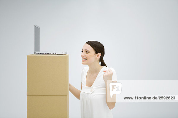 Frau lächelt mit erhobenen Händen und schaut auf einen Laptop  der hoch auf einem Karton steht.