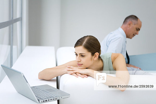 Young woman relaxing on sofa  using laptop computer  businessman reading in background