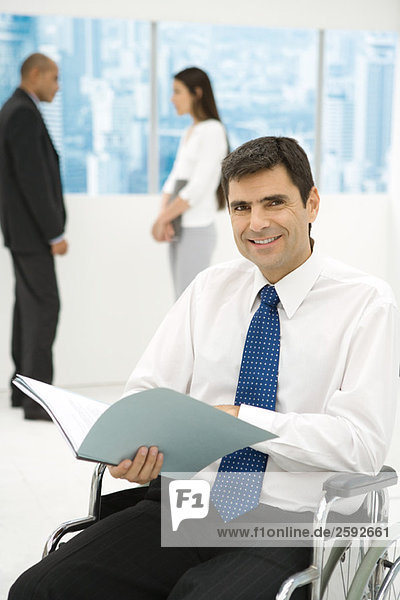 Professional man in wheelchair holding documents  smiling at camera  colleagues in background