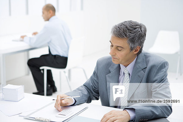 Professional man sitting at desk  using calculator  colleague in background