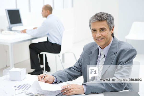 Businessman working at desk  smiling at camera  colleague in background