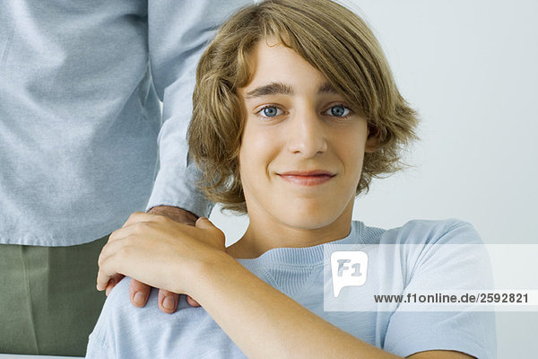 Jugendlicher Junge hält Vaters Hand auf der Schulter  lächelt in die Kamera  Blick in den Ausschnitt