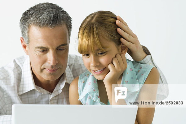 Vater und Tochter schauen zusammen auf den Laptop  die Hand des Mannes auf dem Kopf des Mädchens.