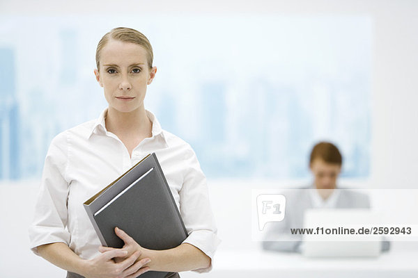 Businesswoman holding folder  looking at camera  male colleague in background