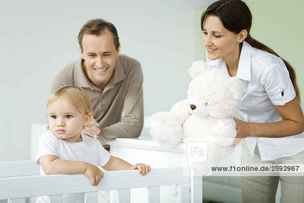 Parents standing beside toddler in crib  mother holding teddy bear  toddler looking away