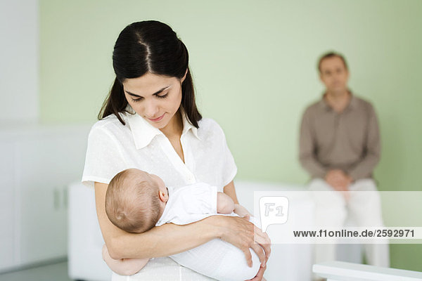 Woman holding sleeping baby  husband sitting in background