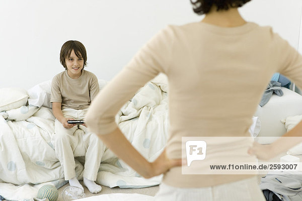 Junge spielt Handheld-Videospiel im unordentlichen Schlafzimmer  Mutter steht im Vordergrund mit Händen auf den Hüften.