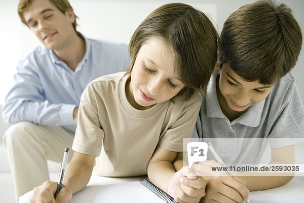 Zwei Jungen schreiben zusammen in ein Notizbuch  Vater sieht im Hintergrund zu.