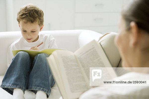 Junge sitzt auf Sofa  schreibt im Buch  Mutter liest im Vordergrund