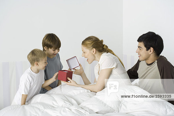 Two boys giving mother present in bed  husband watching