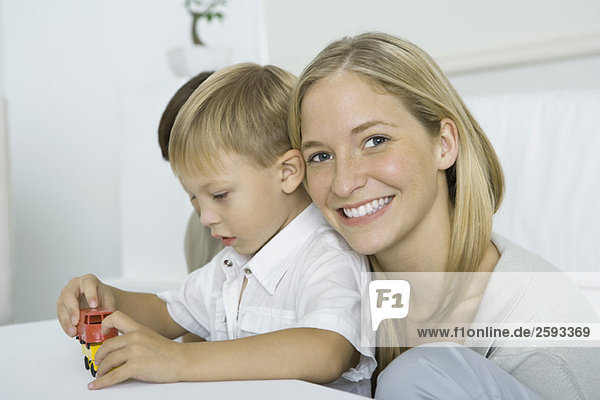 Mother resting her head on son's shoulder  smiling at camera  boy playing with toy cars