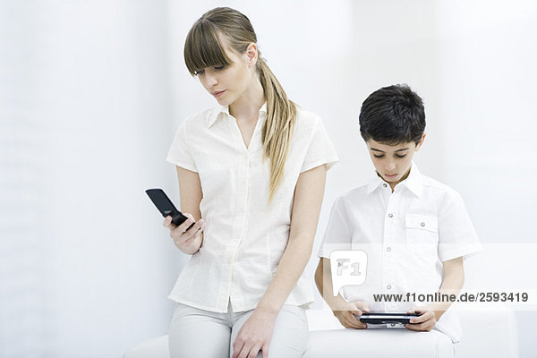 Junge Frau schaut auf das Handy  während der Junge ein Handheld-Videospiel spielt.