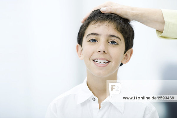 Hand des Erwachsenen auf dem Kopf des Kindes  Junge lächelnd vor der Kamera  abgeschnittene Ansicht