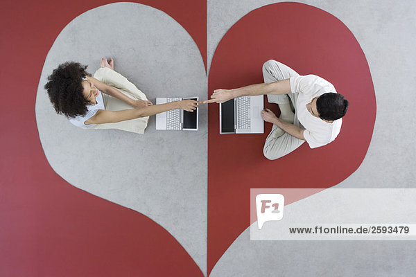 Couple sitting face to face with laptop computers on heart shape  touching fingers  overhead view
