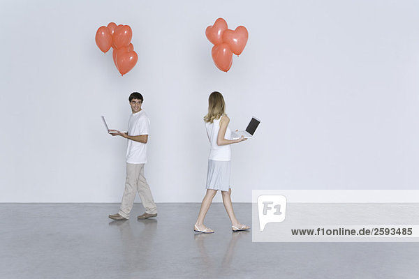 Man and woman walking past each other  both carrying laptop computers and heart balloons