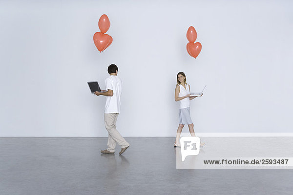 Mann und Frau tragen Laptop-Computer und Herzballons und lächeln sich gegenseitig über die Schultern.
